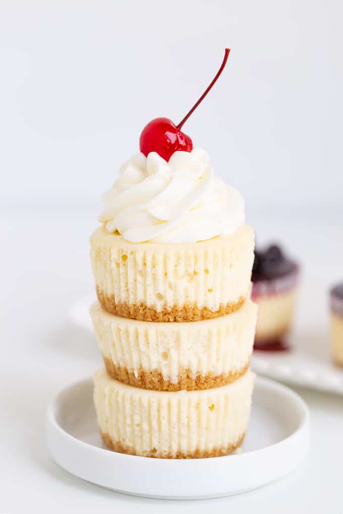 Mini cheesecakes stacked on a small rimmed white plate on a white surface with whipped cream and a cherry on top.