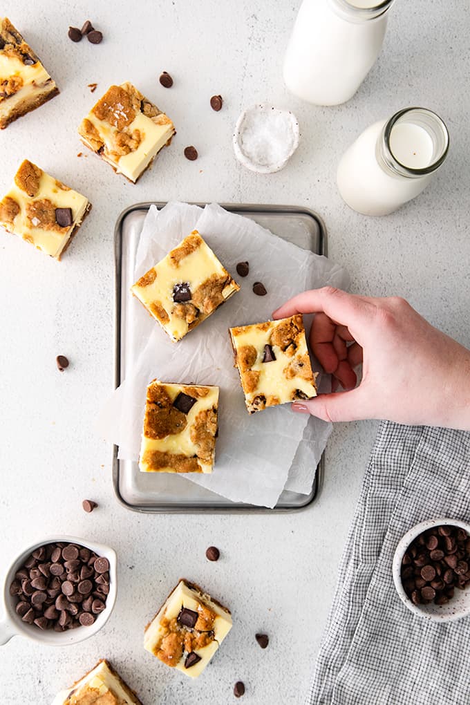 a hand holding a cheesecake bar over top of a pan of bars on a gray surface with chocolate chips and a glass of milk