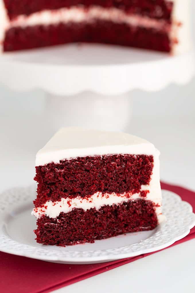 slice of cake on a white plate with a red linen under it and with a white cake plate behind it