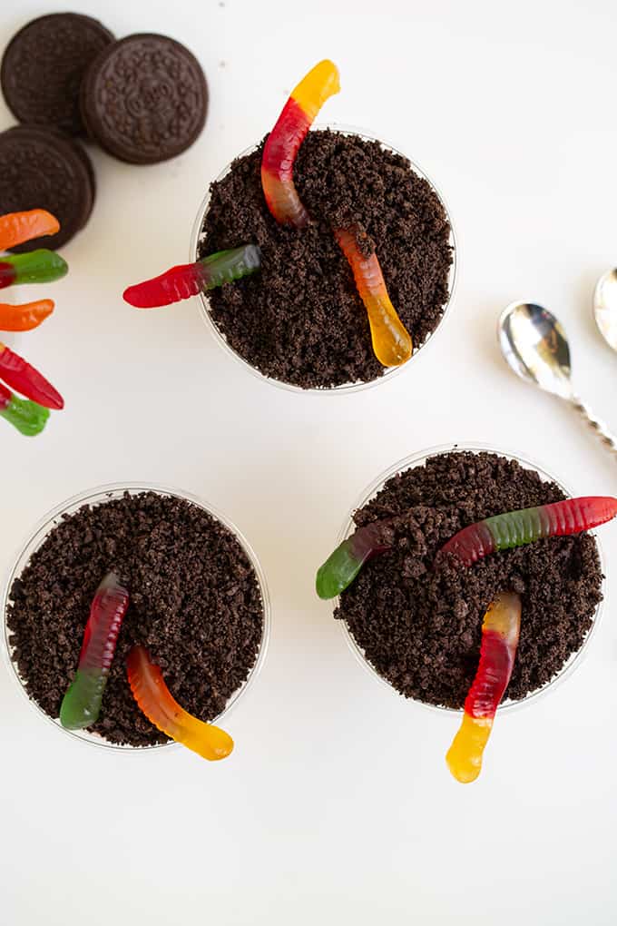 overhead image of cups full of crushed oreos and gummy worms on a white surface with Oreo cookies and a spoon