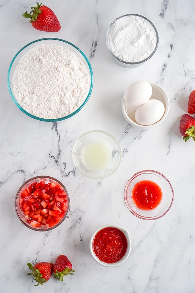overhead photo of ingredients to make cake mix strawberry brownies all laid out in glass bowls on a marble surface