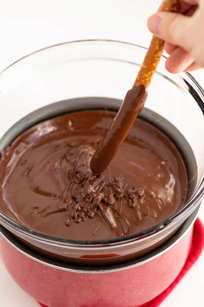 bowl of melted chocolate on a red saucepan with a pretzel rod in hand