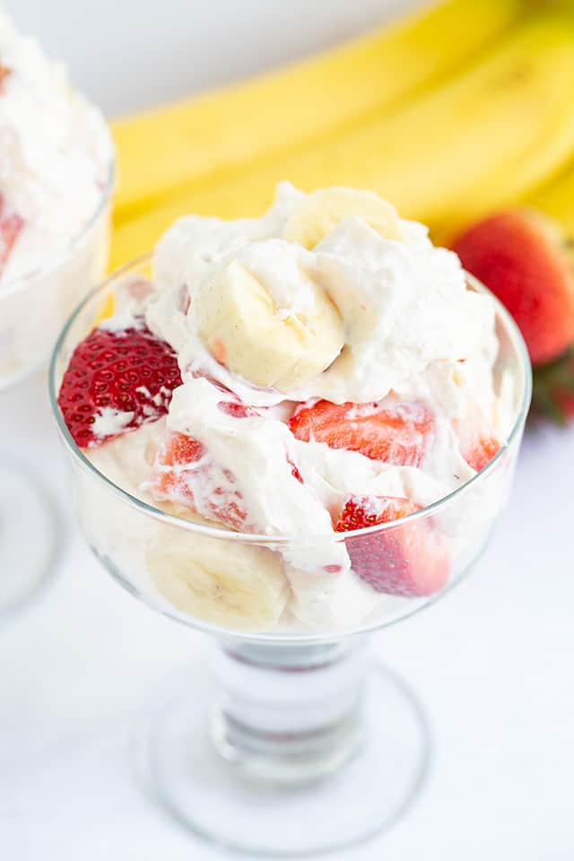 dessert salad in a glass on a white background with banana and strawberries behind it