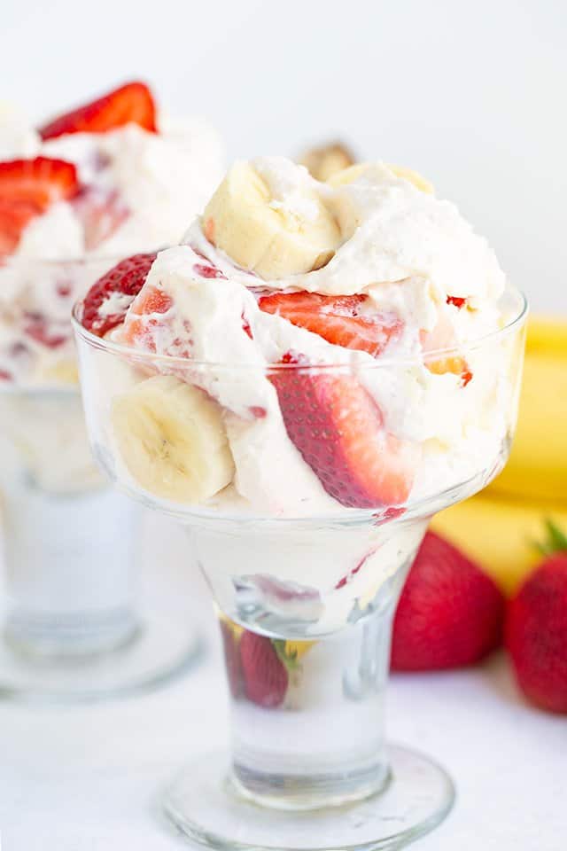 dessert glass filled with cheesecake, fresh strawberries, bananas with fruit behind it