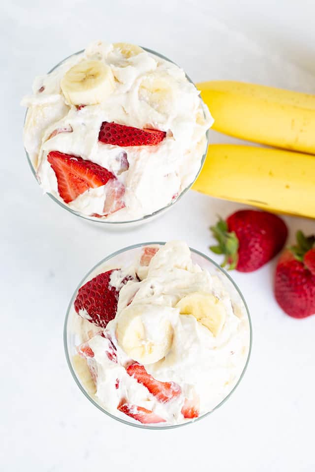 overhead photo of cheesecake salad in two glasses with bananas and strawberries beside it