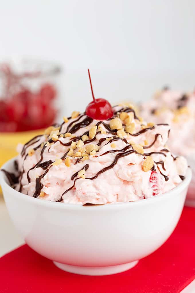 white bowl of dessert salad on a red fabric with bananas and cherries behind it on a white surface