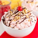 square image showing a close up of the fluff salad with peanuts, chocolate syrup, and a cherry on top on a red fabric