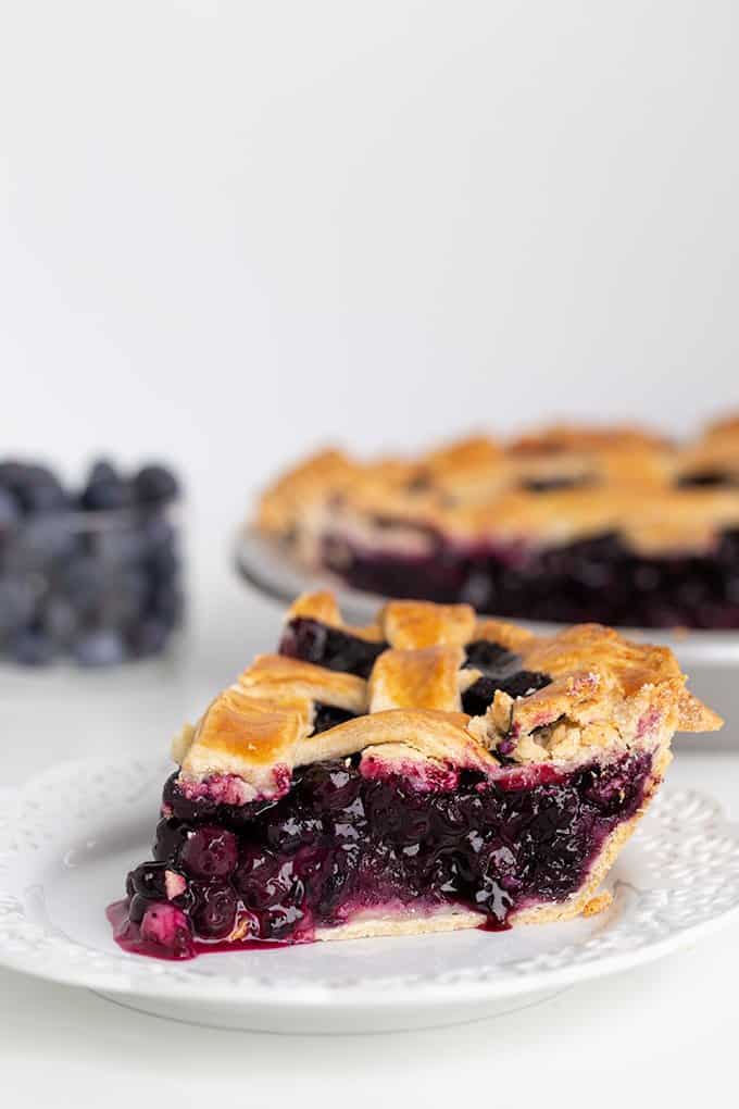 blueberry pie on a white dessert plate with the pie and fresh blueberries behind it on a white surface