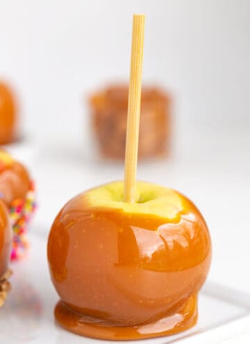 caramel apple on a stick on a white plate on a white surface with caramels behind it