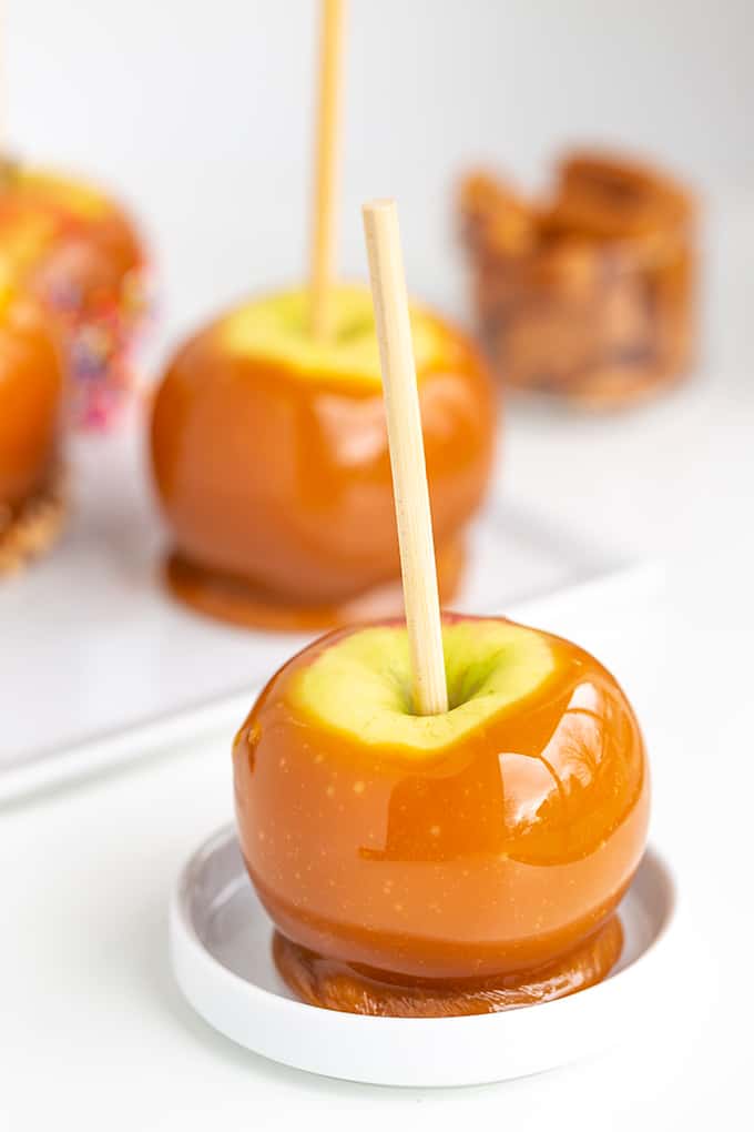 caramel apple on a white round plate on a white surface with a platter of caramel apples behind it