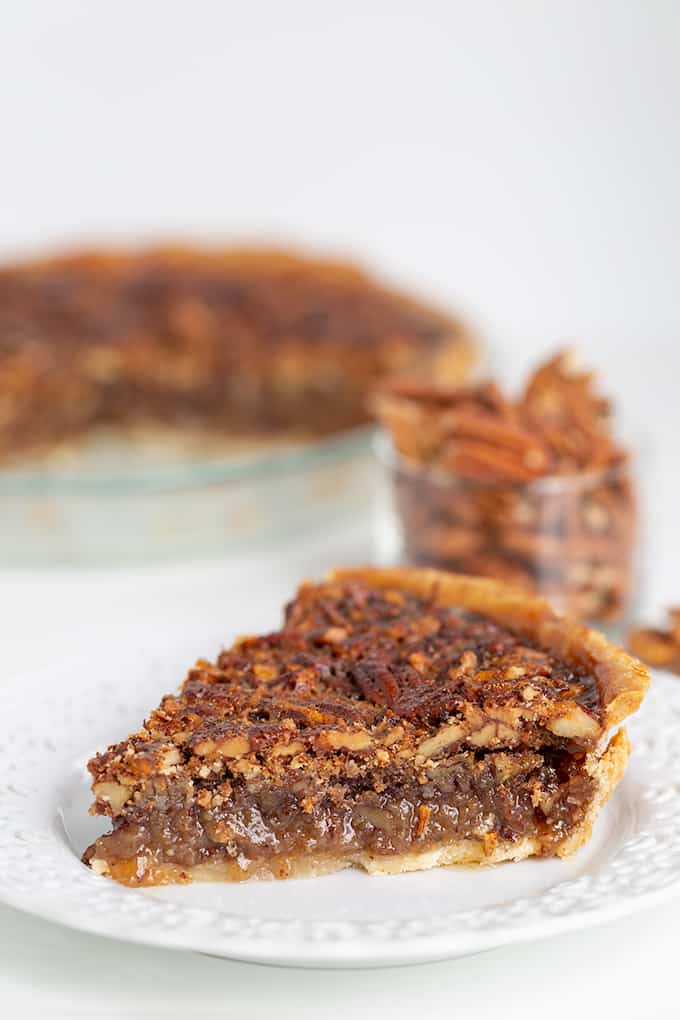 slice of pecan pie on a white lace plate with a small bowl of pecans in a glass bowl on a white surface