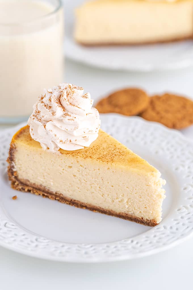 slice of cheesecake on a white lace plate with cookies and eggnog behind the cheesecake on a white surface