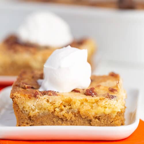 pumpkin bars on a square white plate with whipped cream on top and an orange linen under the plate