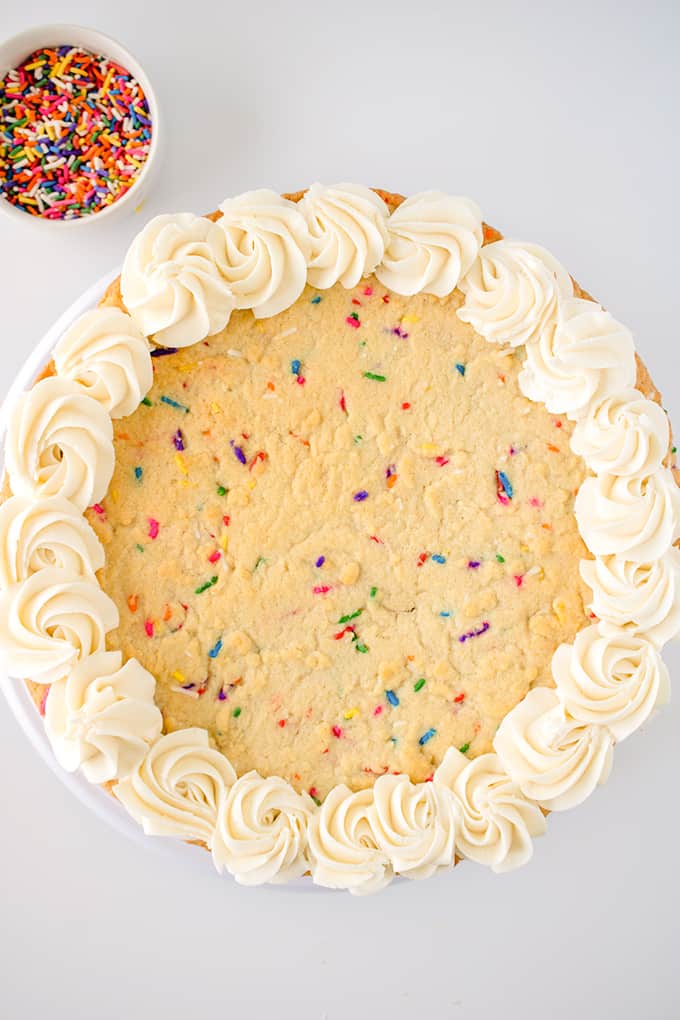homemade cookie cake on a white surface with rainbow sprinkles in a white bowl next to it