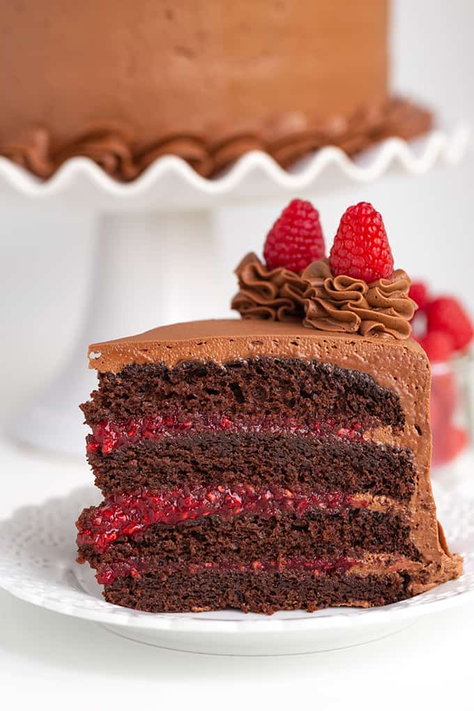 slice of cake on a white plate with the rest of the chocolate raspberry cake on a white cake stand behind the slice of cake