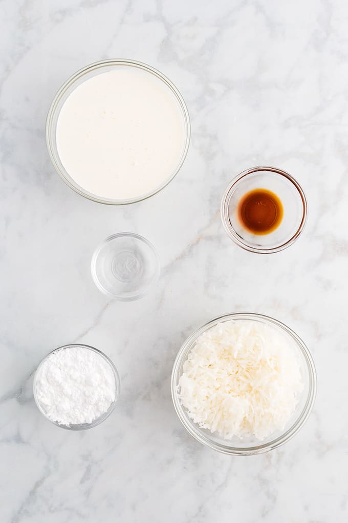 Frosting ingredients: heavy whipping cream, vanilla extract, coconut extract, powdered sugar, coconut in bowls on a marble background