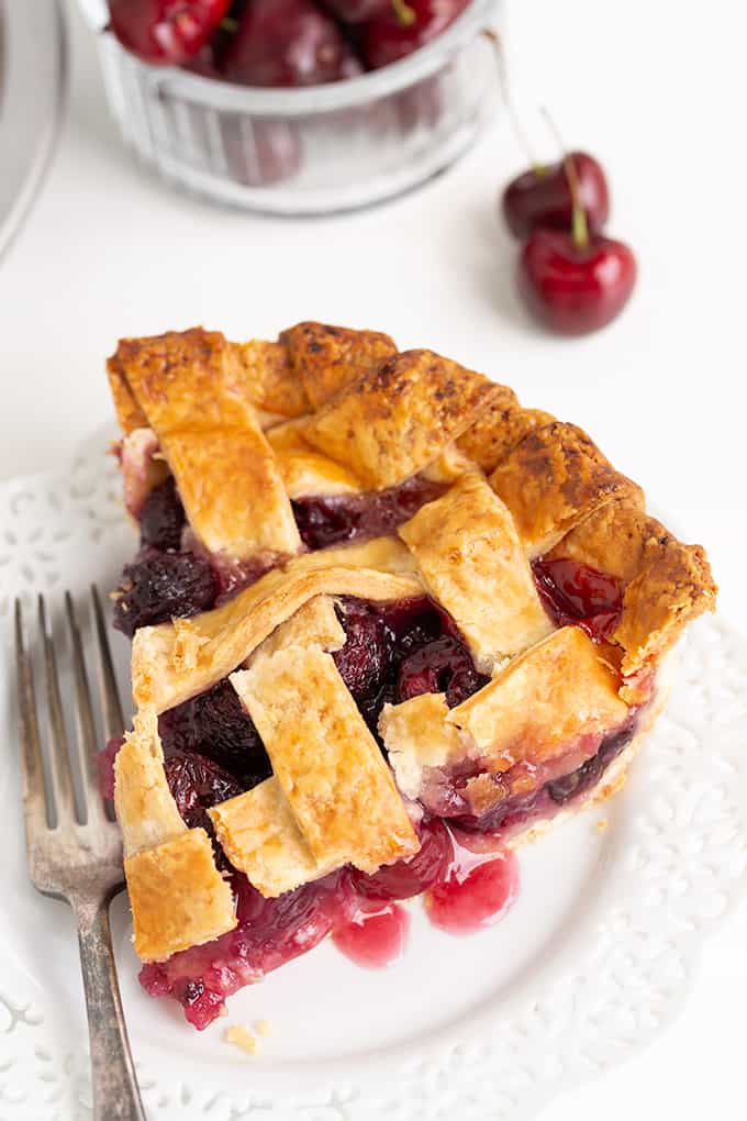 cherry pie on a white lace plate with a fork on the plate and fresh cherries beside the plate