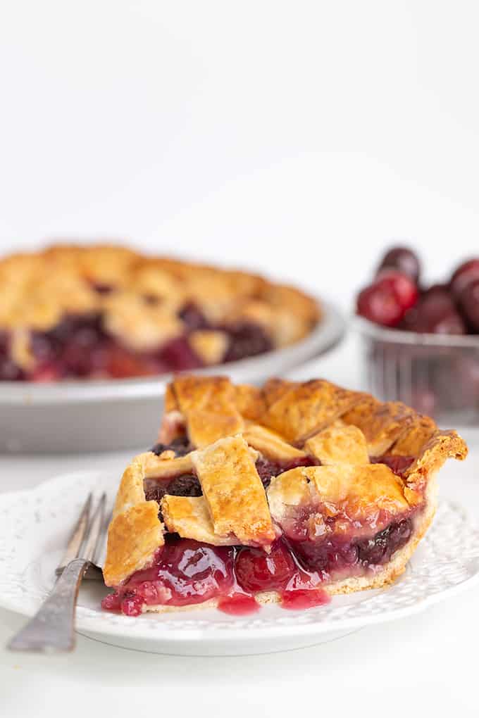 close up of the side of a cherry pie showing the juice leaking out onto the white plate with a fork on the plate, a bowl of cherries behind the slice of pie, and the pie plate beside the cherries