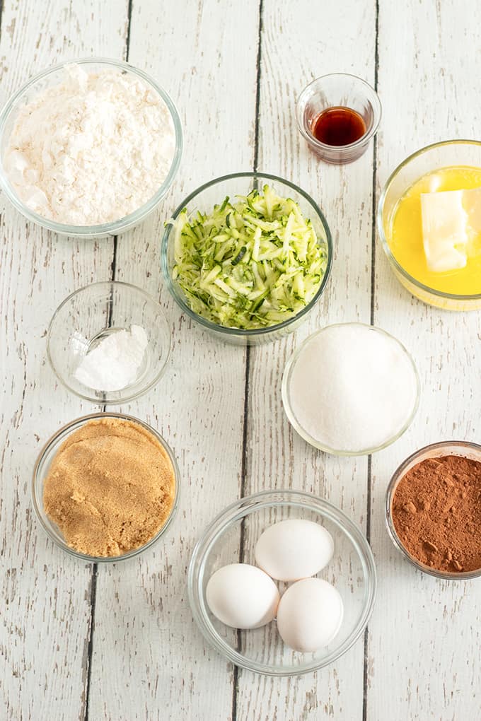 Overhead photo of Ingredients: flour, cocoa, baking soda, baking powder, salt, eggs, granulated sugar, light brown sugar, shredded zucchini, butter, and vanilla extract in glass bowls on a white planked blackground