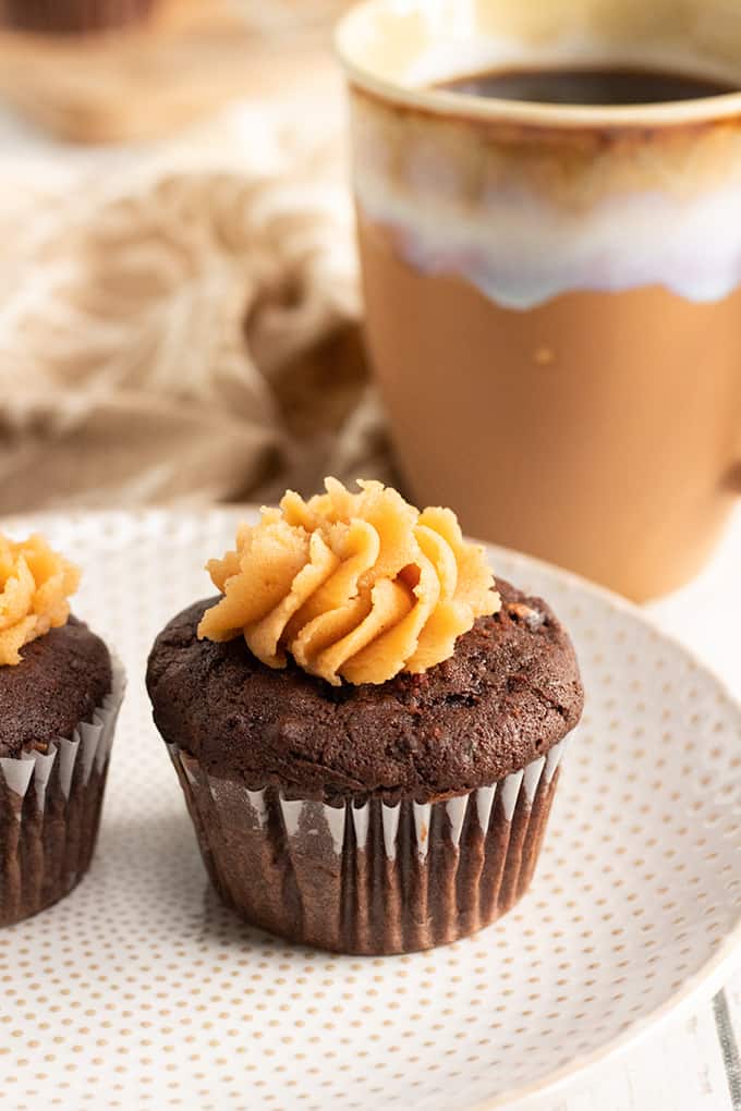 Close up side view of chocolate zucchini cupcake with peanut butter frosting on a plate and coffee in a brown mug in the background.