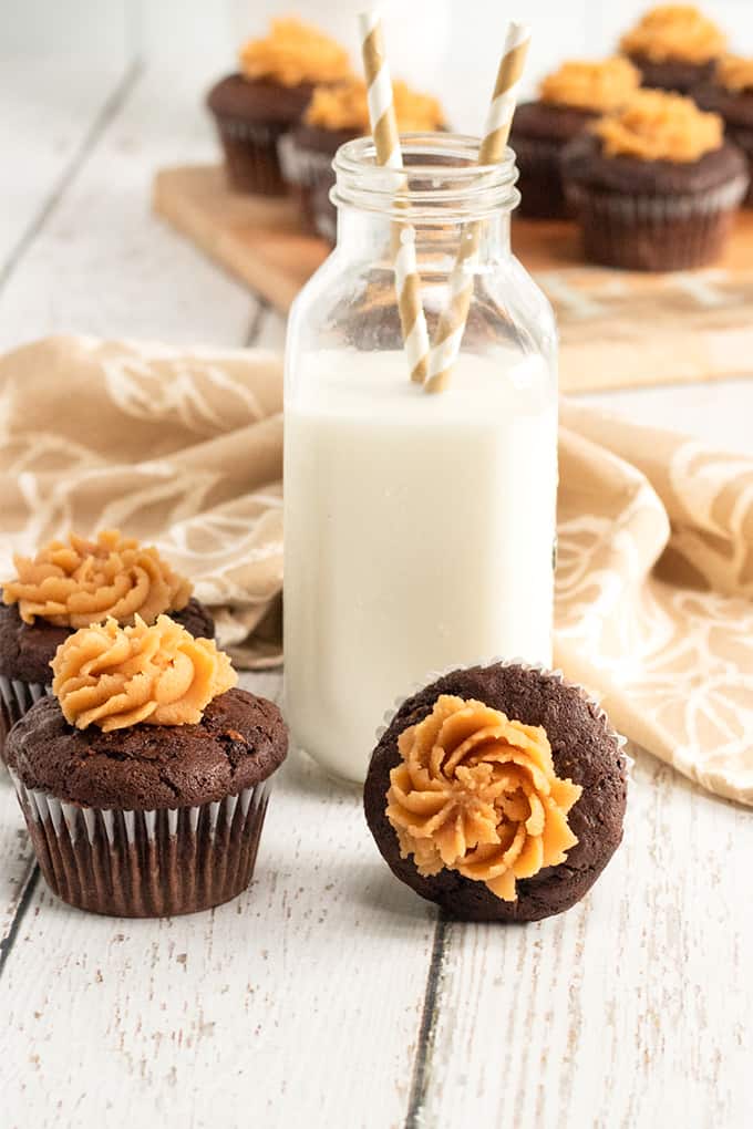 More distant view of several chocolate zucchini cupcakes on white wood with a bottle of milk and a beige linen behind the bottle