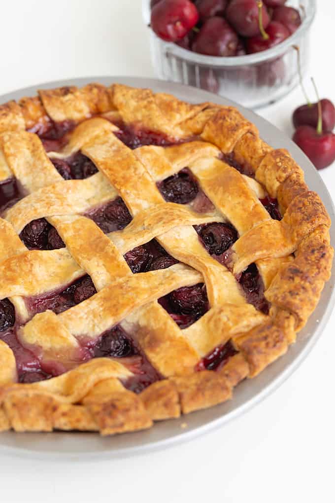 lattice pie crust cherry pie in a plan with a bowl of fresh cherries behind it sitting on a white surface