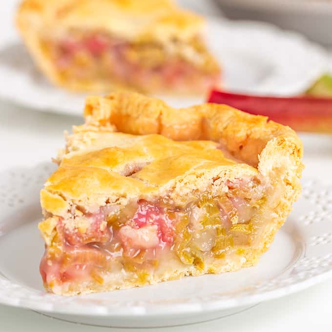 square image of a slice of rhubarb pie on a white plate with a stalk of rhubarb behind the slice of pie