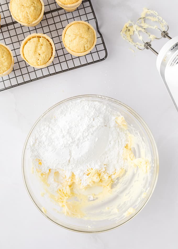 Powdered sugar and butter in a mixing bowl for buttercream frosting. Baked cupcakes on a cooling rack.