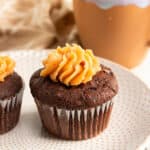 chocolate zucchini cupcake with peanut butter frosting in a paper cupcake liner on a white textured plate with a brown coffee cup behind it