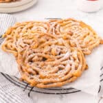 Three homemade funnel cakes on a cooling rack with powdered sugar.