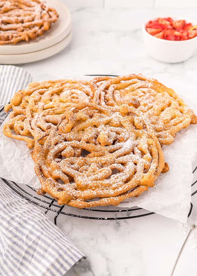 Three homemade funnel cakes on a cooling rack with powdered sugar.