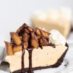 A slice of Peanut Butter Pie on a white dessert plate with a pile of peanut butter cups and a drizzle of chocolate syrup