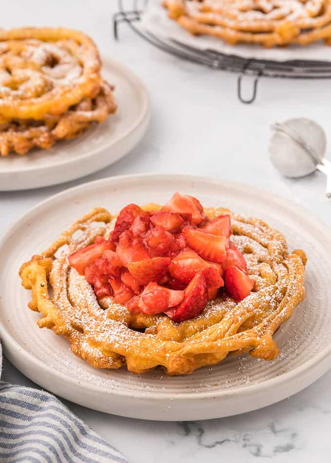 Homemade Funnel Cakes topped with powdered sugar and strawberries on a white plate on a marble surface with a blue striped linen next to it