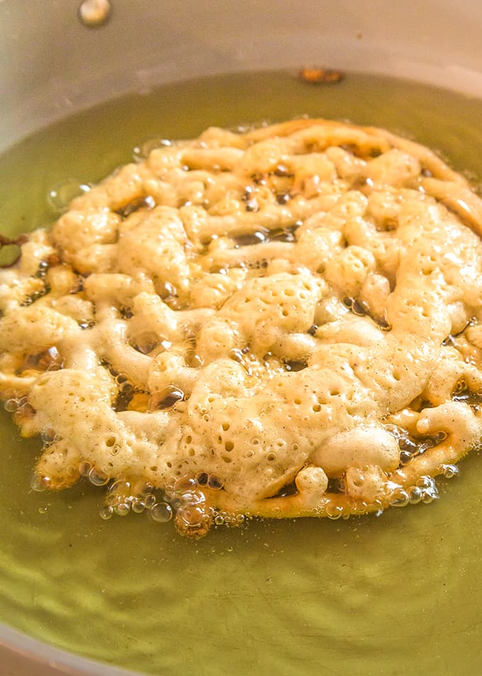 A homemade funnel cake is being cooked in a frying pan with canola oil