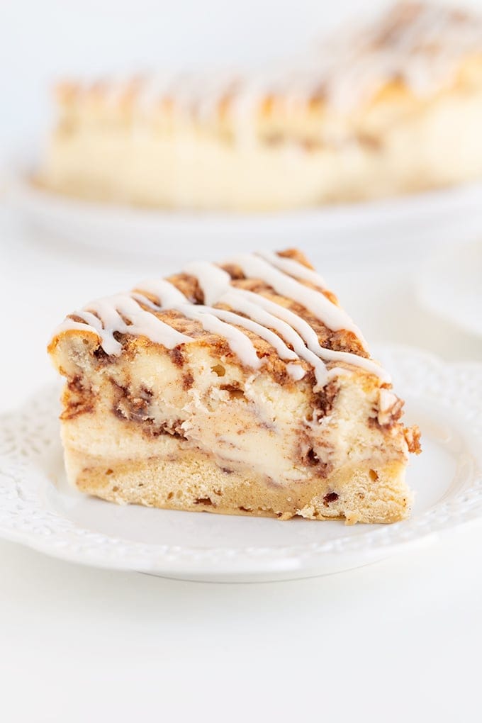 A slice of cinnamon swirl cheesecake on a white plate with the remaining cheesecake behind on on a white background