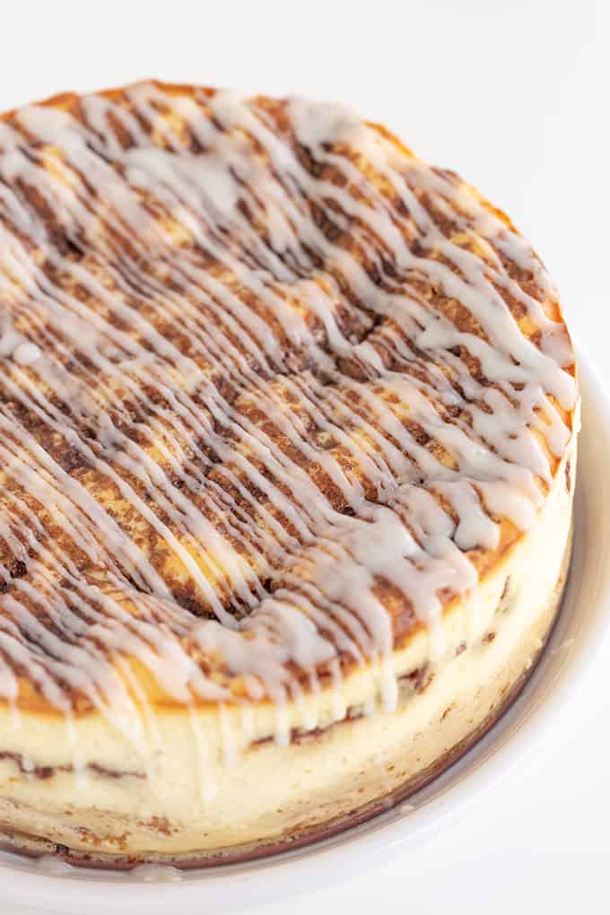 A Cinnamon Roll Cheesecake with icing on a rimmed white plate with a white background