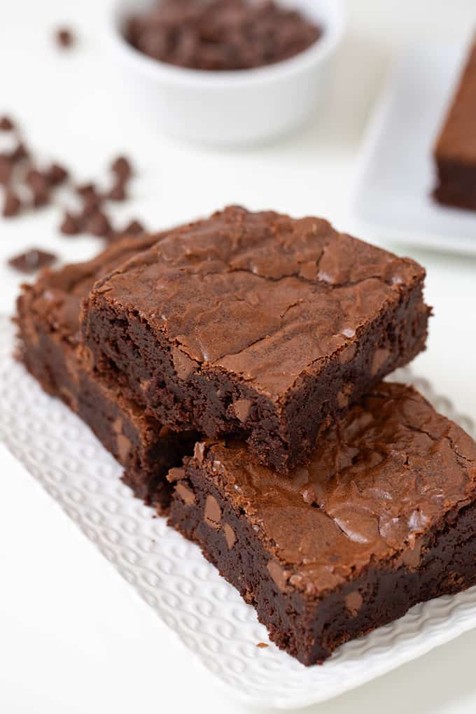 Improved chocolate brownies presented on a rectangle white plate with chocolate chips scattered on the white background