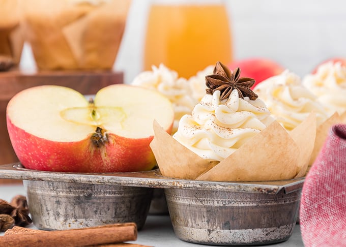 A tray of apple cider cupcakes with whipped cream and cinnamon sticks.