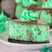A slice of Mint Chocolate Cheesecake with a chocolate cookie crust, cool green filling, chocolate pieces, a swirl of minty green cream, and a square of chocolate on top.