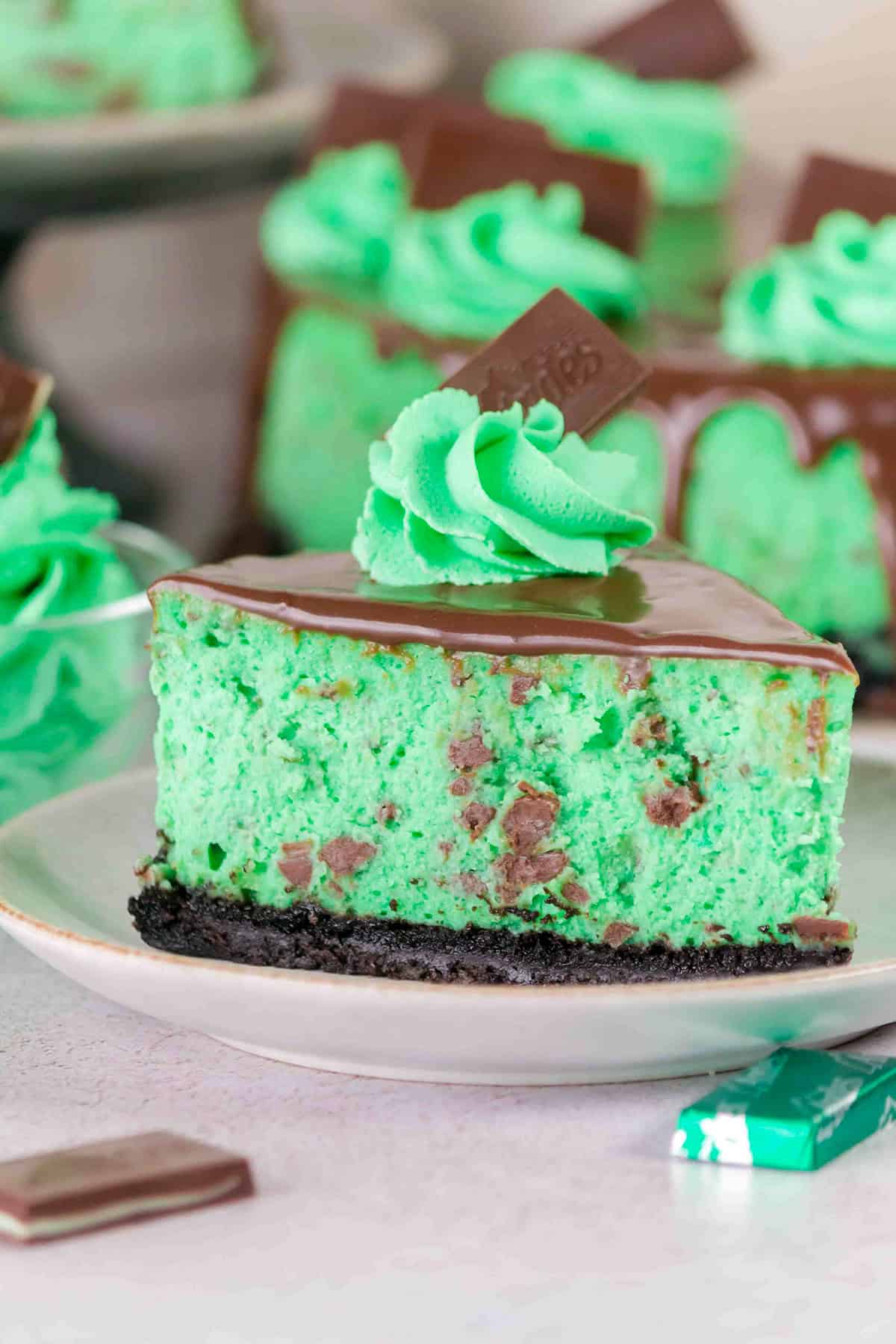 A slice of Mint Chocolate Cheesecake with a chocolate cookie crust, cool green filling, chocolate pieces, a swirl of minty green cream, and a square of chocolate on top.