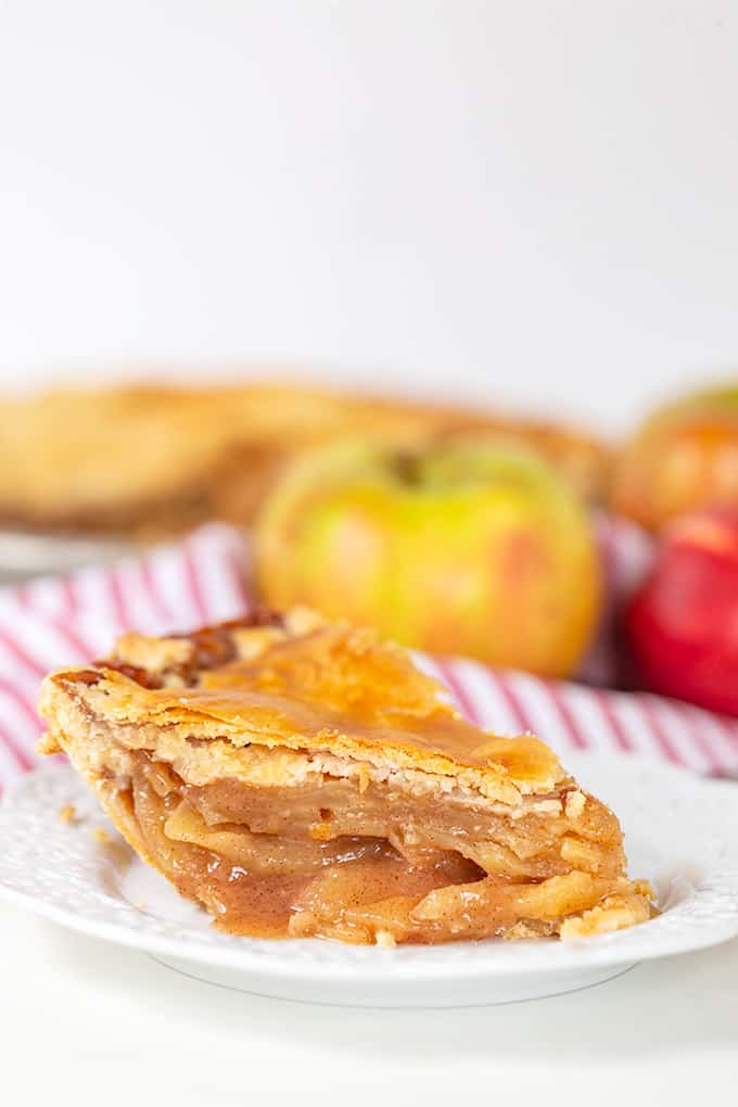 A slice of apple pie on a white plate with red and white striped linen with apples behind it