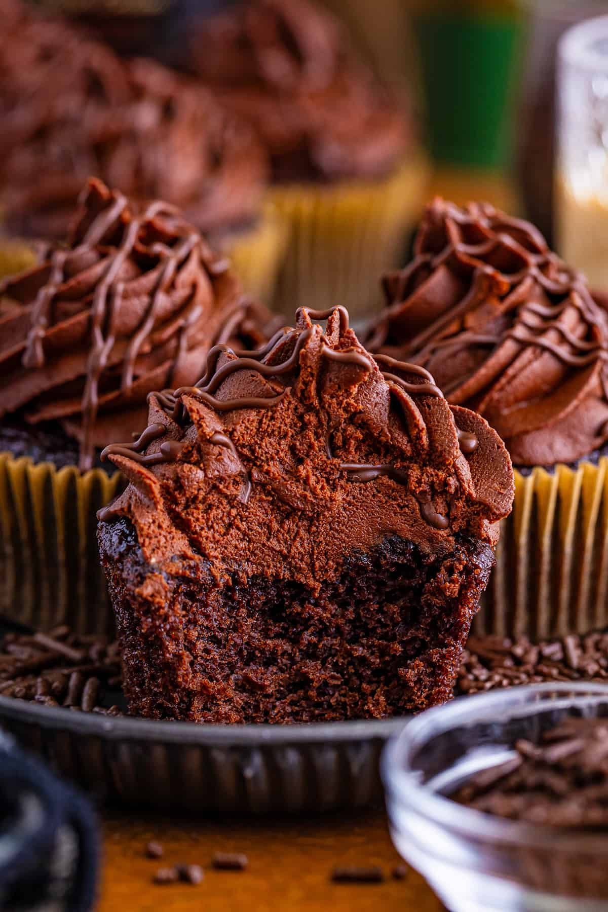 Chocolate cupcakes with swirls of Baileys frosting on a wooden surface, surrounded by dessert ingredients.