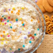 A bowl of coconut cream dessert dip with colorful sprinkles, served with an assortment of cookies and pretzels.