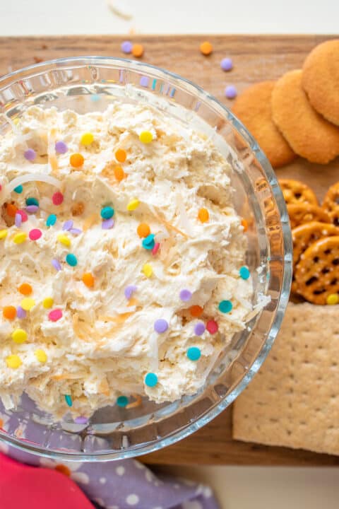 A bowl of coconut cream dessert dip with colorful sprinkles, served with an assortment of cookies and pretzels.