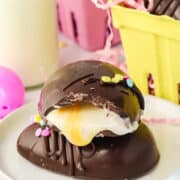 A chocolate dessert with a glossy exterior and a creamy filling, mimicking Copycat Creme Egg, topped with sprinkles, presented on a plate with an Easter candy-inspired festive background.
