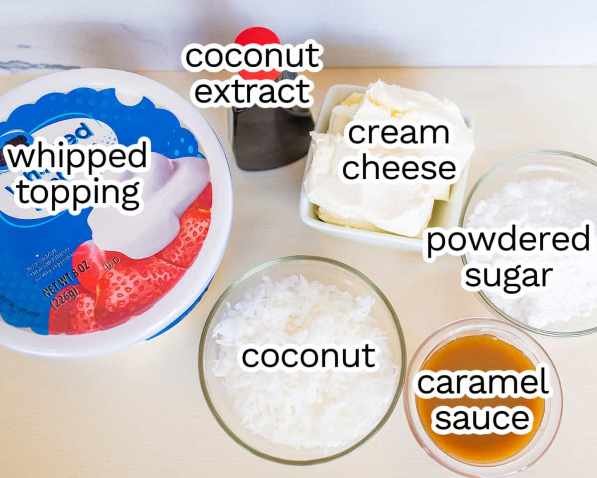 Ingredients for a Coconut Cream Dip dessert recipe including whipped topping, coconut extract, cream cheese, powdered sugar, shredded coconut, and caramel sauce, labeled and arranged on a kitchen counter.