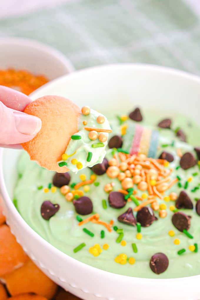 A hand dipping a vanilla wafer into a bowl of St. Patrick's Day Booty Dip, tinted green and topped with sprinkles and chocolate chips.