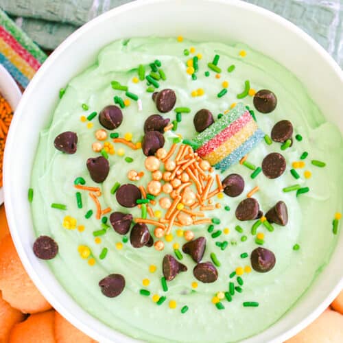 A bowl of green-tinted St. Patrick's Day dessert dip garnished with chocolate chips, sprinkles, and a rainbow candy, served with cookies on the side.
