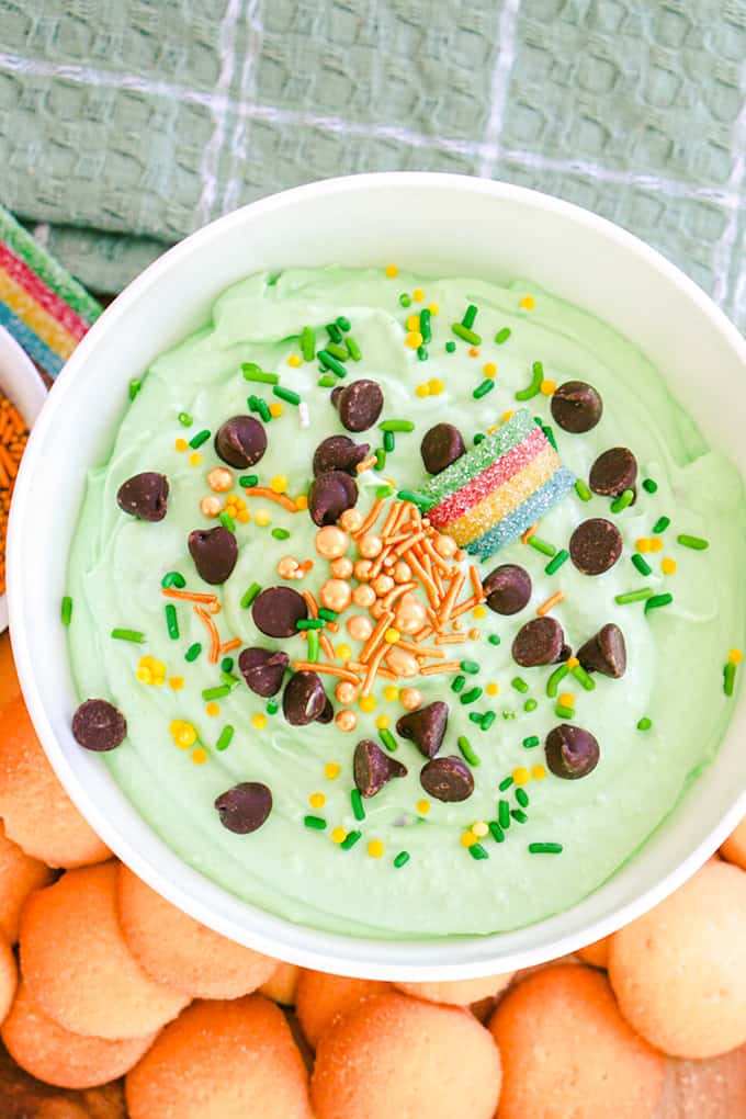 A bowl of green-tinted St. Patrick's Day dessert dip garnished with chocolate chips, sprinkles, and a rainbow candy, served with cookies on the side.