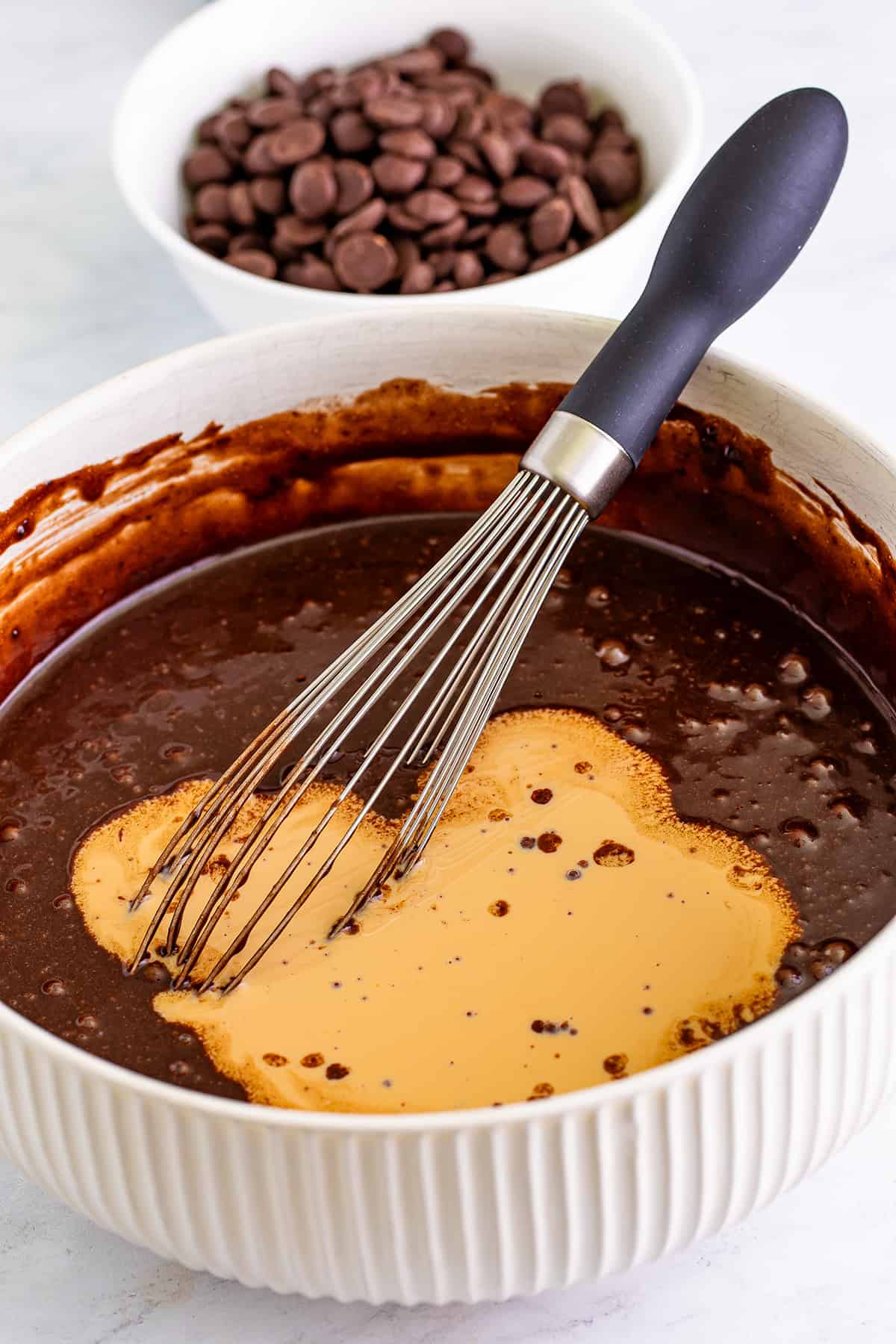 A whisk in a bowl with melted chocolate and a heart-shaped pool of Baileys-flavored cream, with chocolate chips in the background.